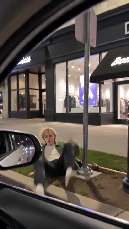Example frame: a young man sitting dazed on a grass strip beside a signpost on a city street at night