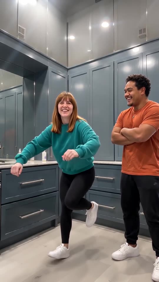 Fotograma de ejemplo: una mujer con sudadera verde azulado zapateando mientras un hombre con camiseta naranja mira con los brazos cruzados en una cocina azul grisácea